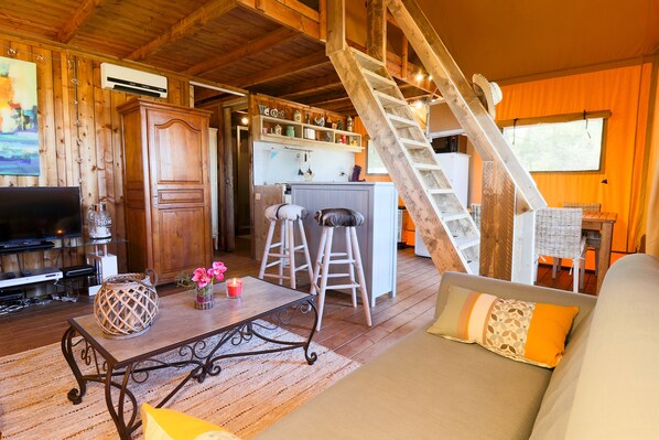 A cozy cabin interior with a wooden floor, a sofa with cushions, a coffee table, a TV, and a kitchen area with a refrigerator and shelves.