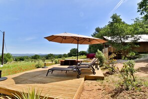 A wooden deck with lounge chairs and an umbrella, surrounded by greenery and a view of the countryside.