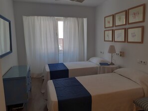 A hotel room with two beds, a desk, a chair, a lamp, and a window with curtains. - Fabulous, Ground floor, Pine Walk apartment with large terrace and beach access (Port de Pollença)