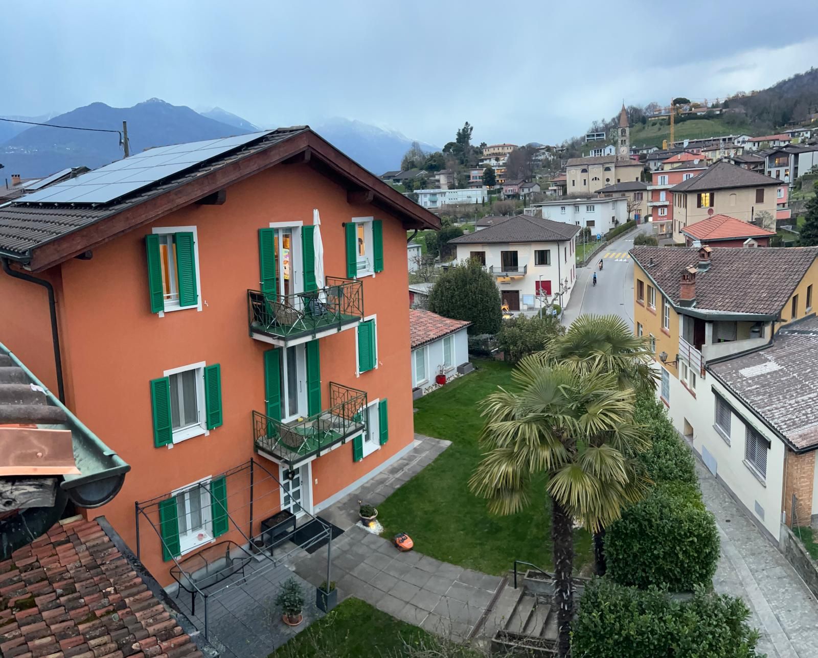 Un immeuble à plusieurs étages doté de balcons, d’un petit jardin et donnant sur la rue, avec d’autres immeubles et des montagnes en arrière-plan.