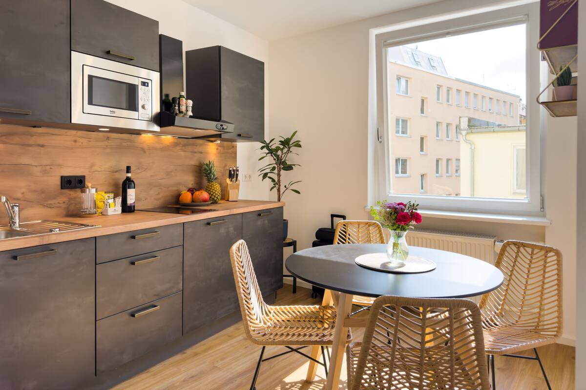 Una cocina moderna con armarios oscuros, un microondas y una encimera de madera. Hay una zona de comedor con sillas de mimbre y una mesa, además de un ventanal con vistas a edificios.