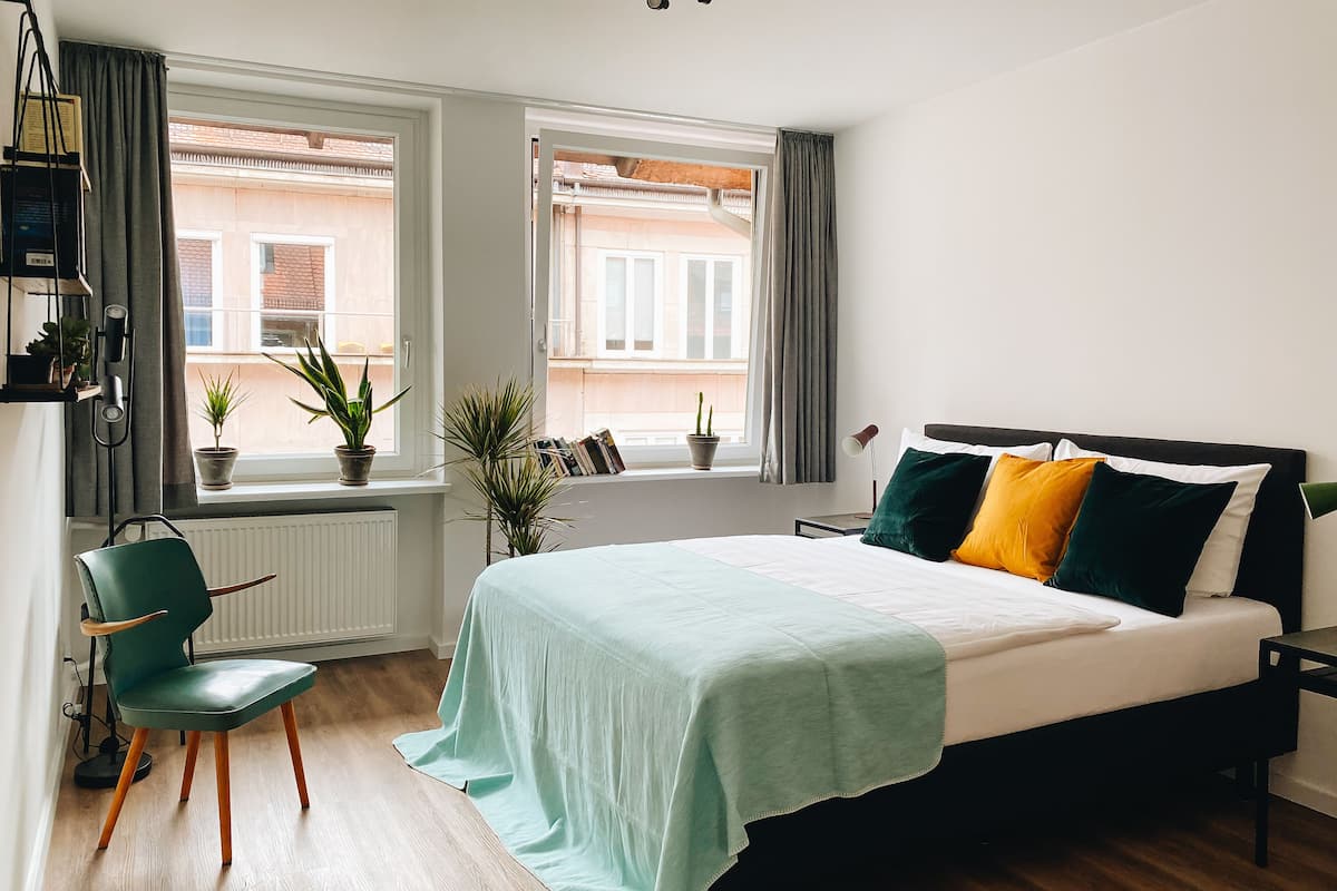 Un dormitorio con cama, una silla, dos ventanas y un radiador.