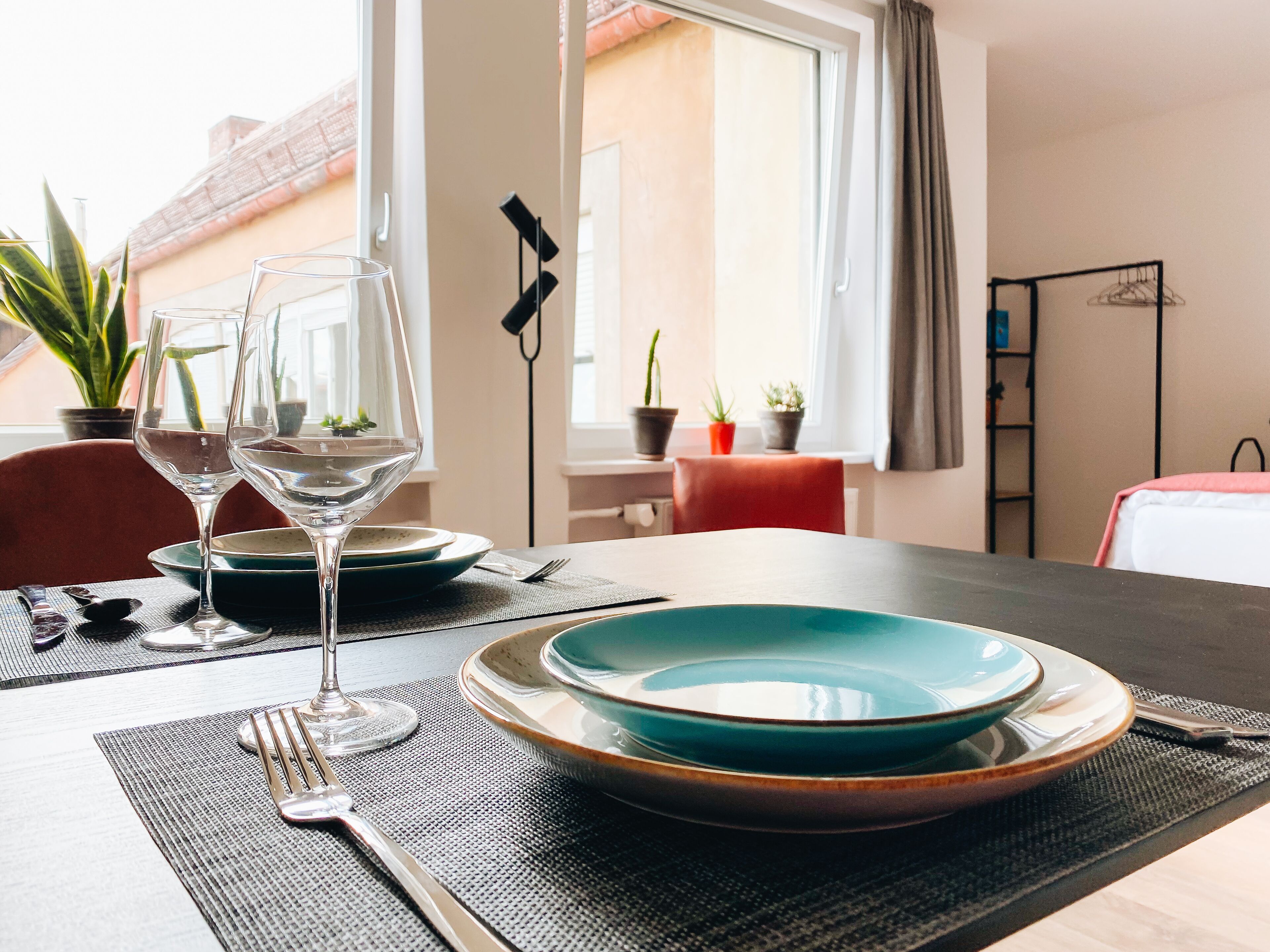 A dining table set with plates, a glass, and cutlery, with a view of a window and potted plants in the background.