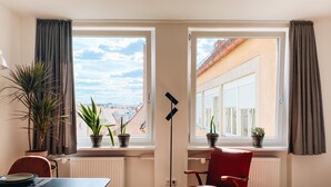 A room with a dining table, a red chair, and two windows with curtains. There are potted plants on the windowsill and a floor lamp.