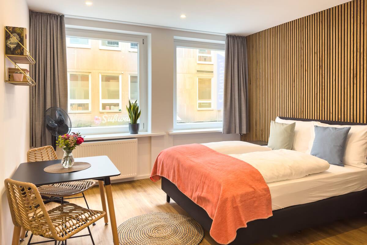 Habitación de hotel moderna con una cama grande, una mesita con sillas, una planta en maceta y vistas de la ciudad a través de amplios ventanales.