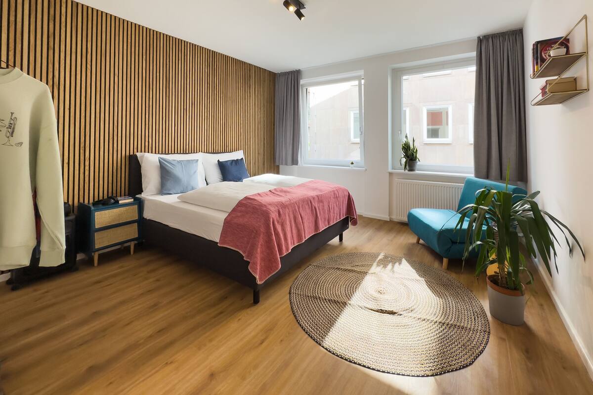 Un dormitorio moderno con una cama, mesitas de noche, un sillón azul, una planta en maceta y vistas al exterior.