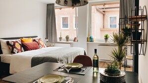 A modern hotel room with a neatly made bed, a dining table set for two, and a view of the city through large windows.