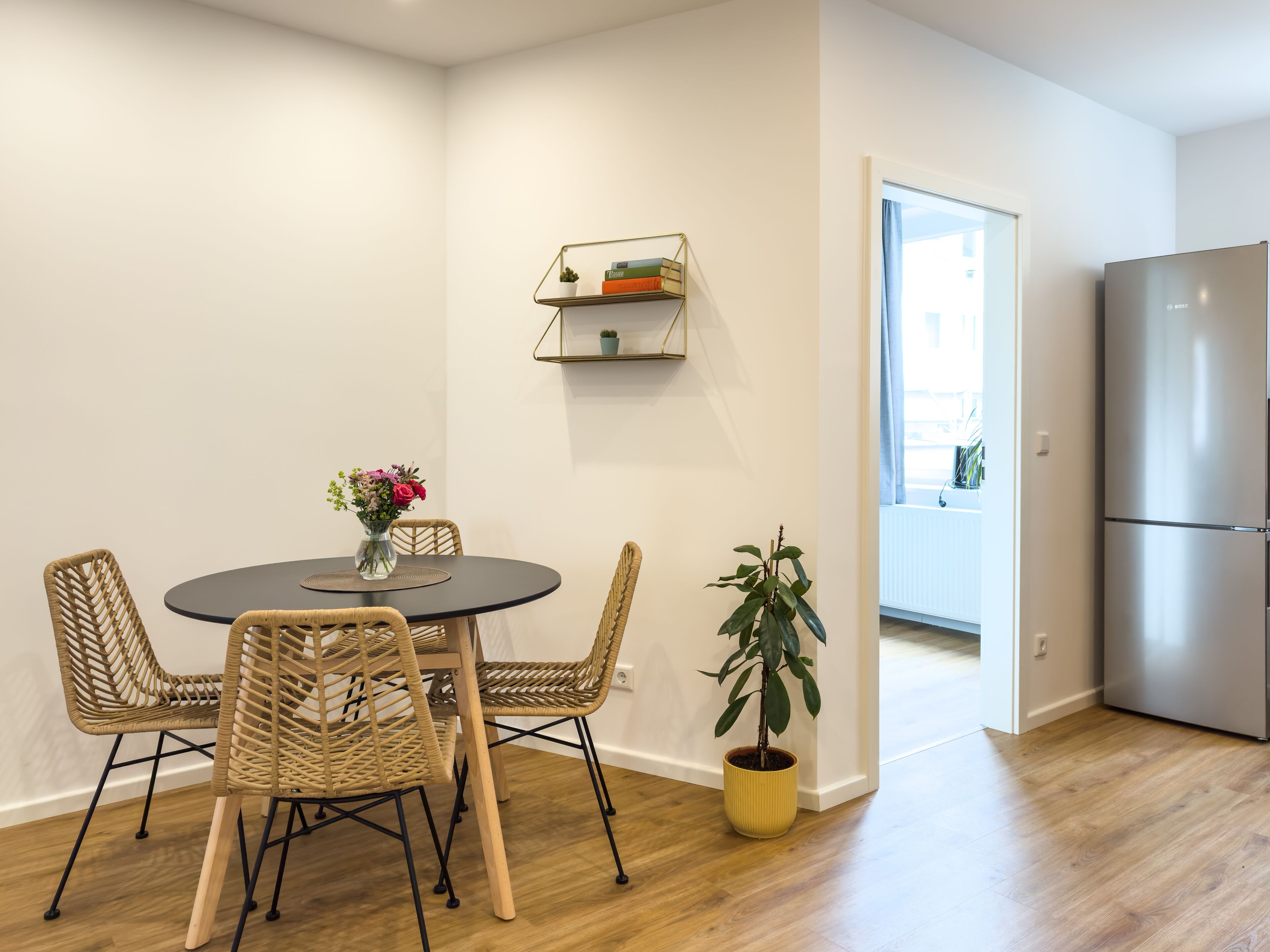 A modern kitchen with a stainless steel refrigerator, a dining area with wicker chairs and a round table, and a potted plant.