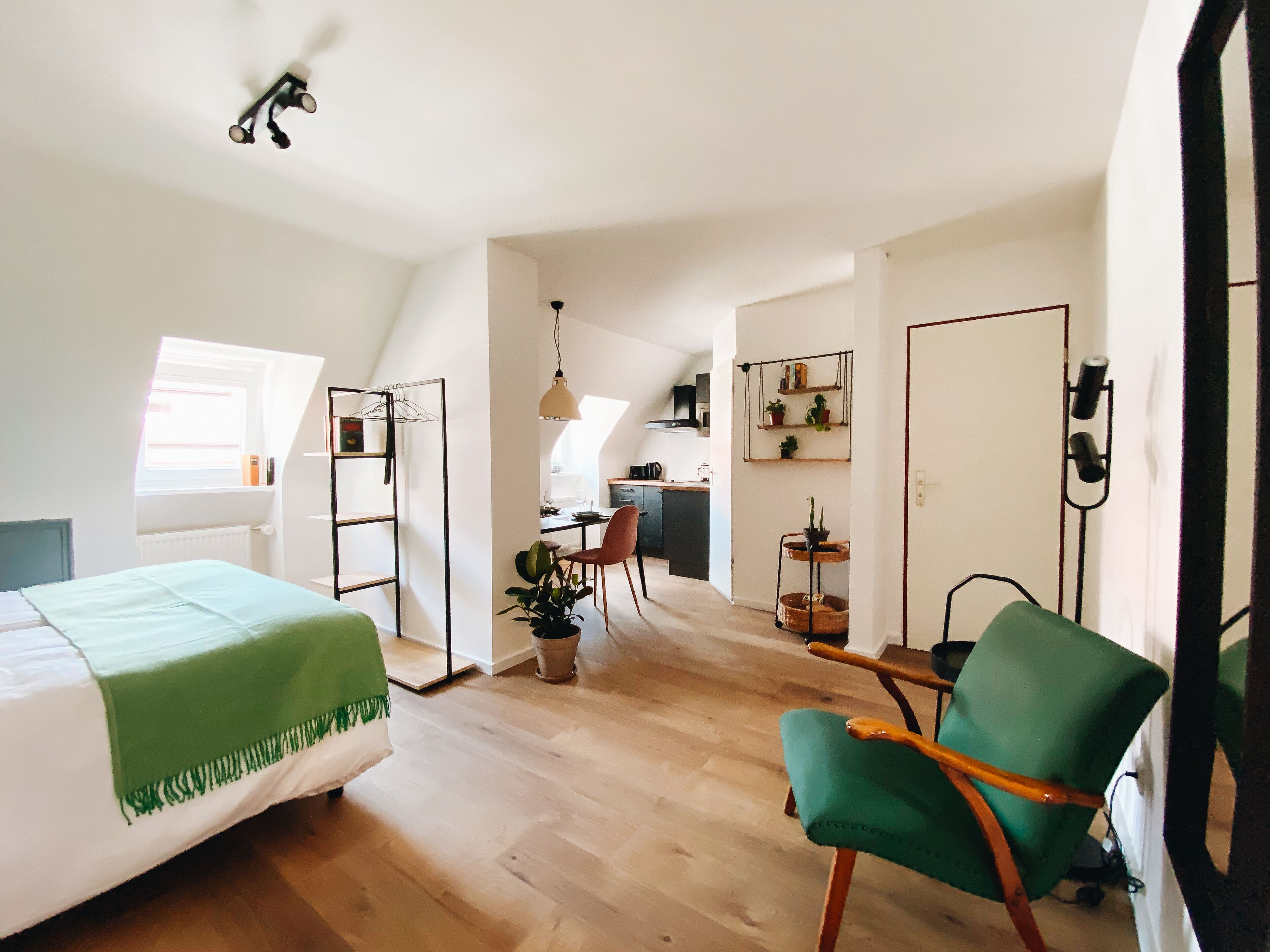 A compact, well-lit room with a bed, a green armchair, a desk, and a kitchenette in the background.