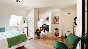 A compact, well-lit room with a bed, a green armchair, a desk, and a kitchenette in the background.