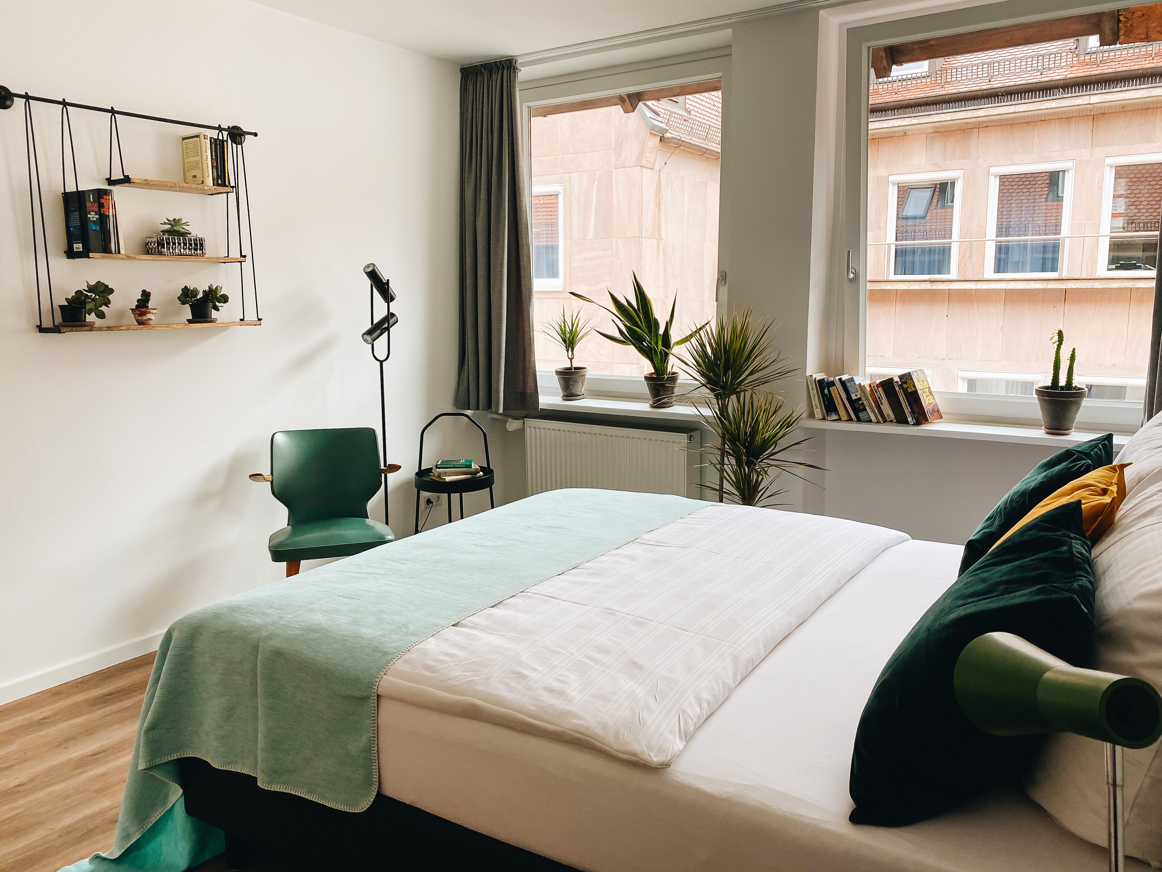 Ein Schlafzimmer mit einem Bett, einem Sessel, einem Regal mit Pflanzen und einem Fenster mit Blick auf Gebäude.