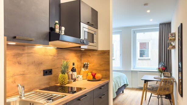 A modern kitchen with a wooden backsplash, stainless steel sink, and a microwave. There is a dining area with a table and chairs, and a window with curtains.