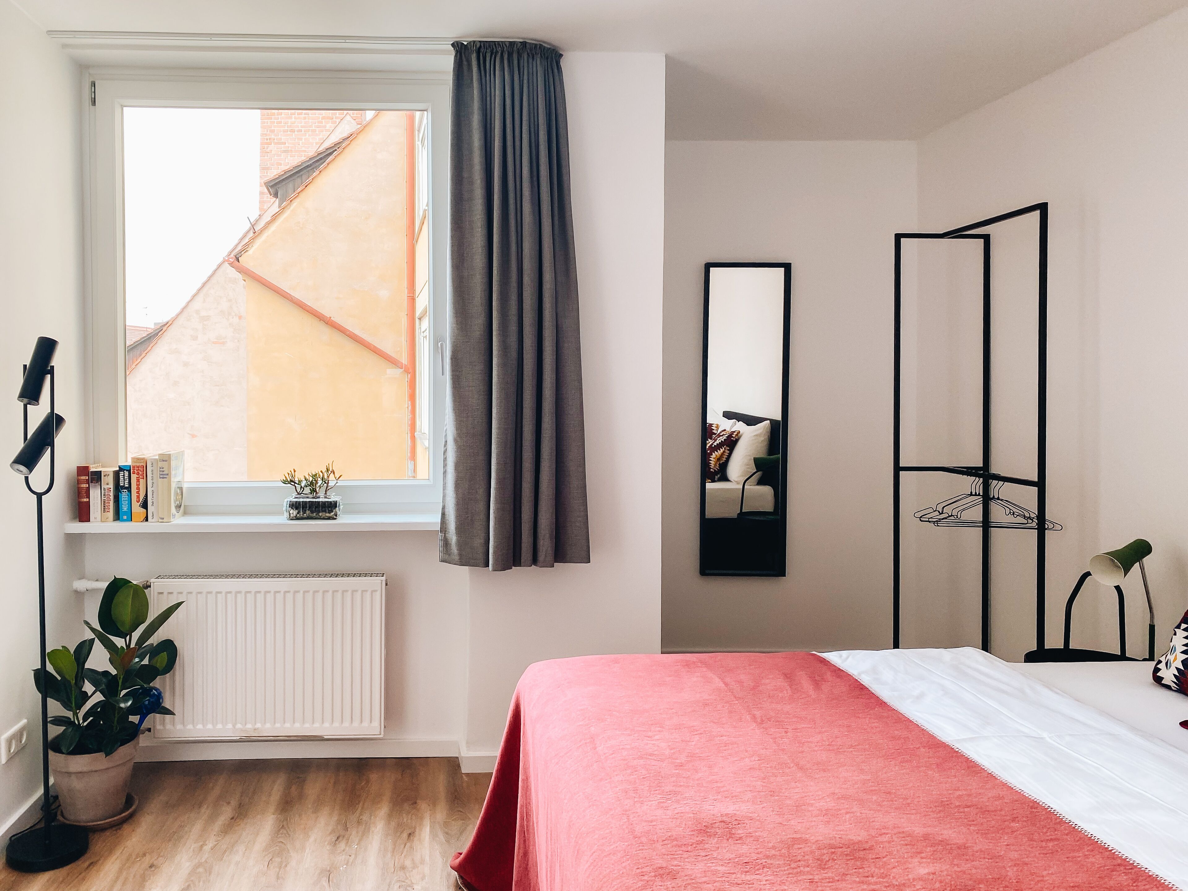 Quarto com cama, janela com vista para edifícios, radiador, planta em vaso e estante de livros.
