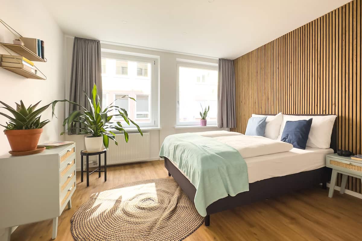 Un dormitorio con cama, mesita de noche, plantas en macetas, cómoda y ventana con cortinas.