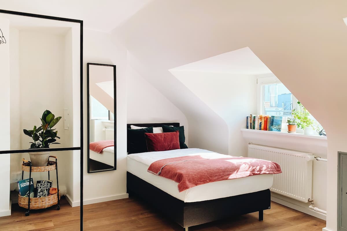 Un dormitorio con cama, espejo, radiador, ventana y una planta.