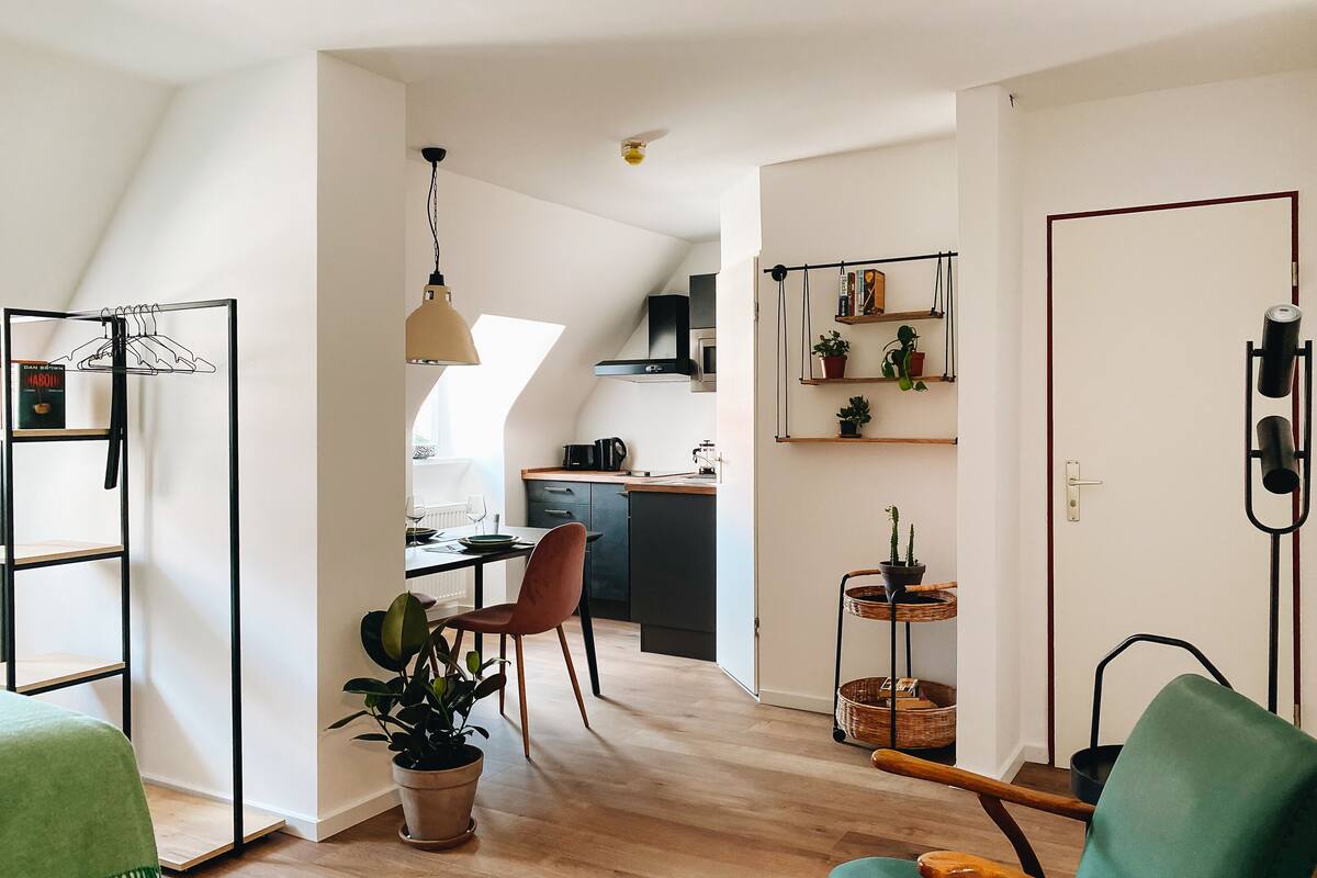 Un apartamento moderno con comedor, cocina y sala de estar que incluye un sillón verde y una planta en maceta.