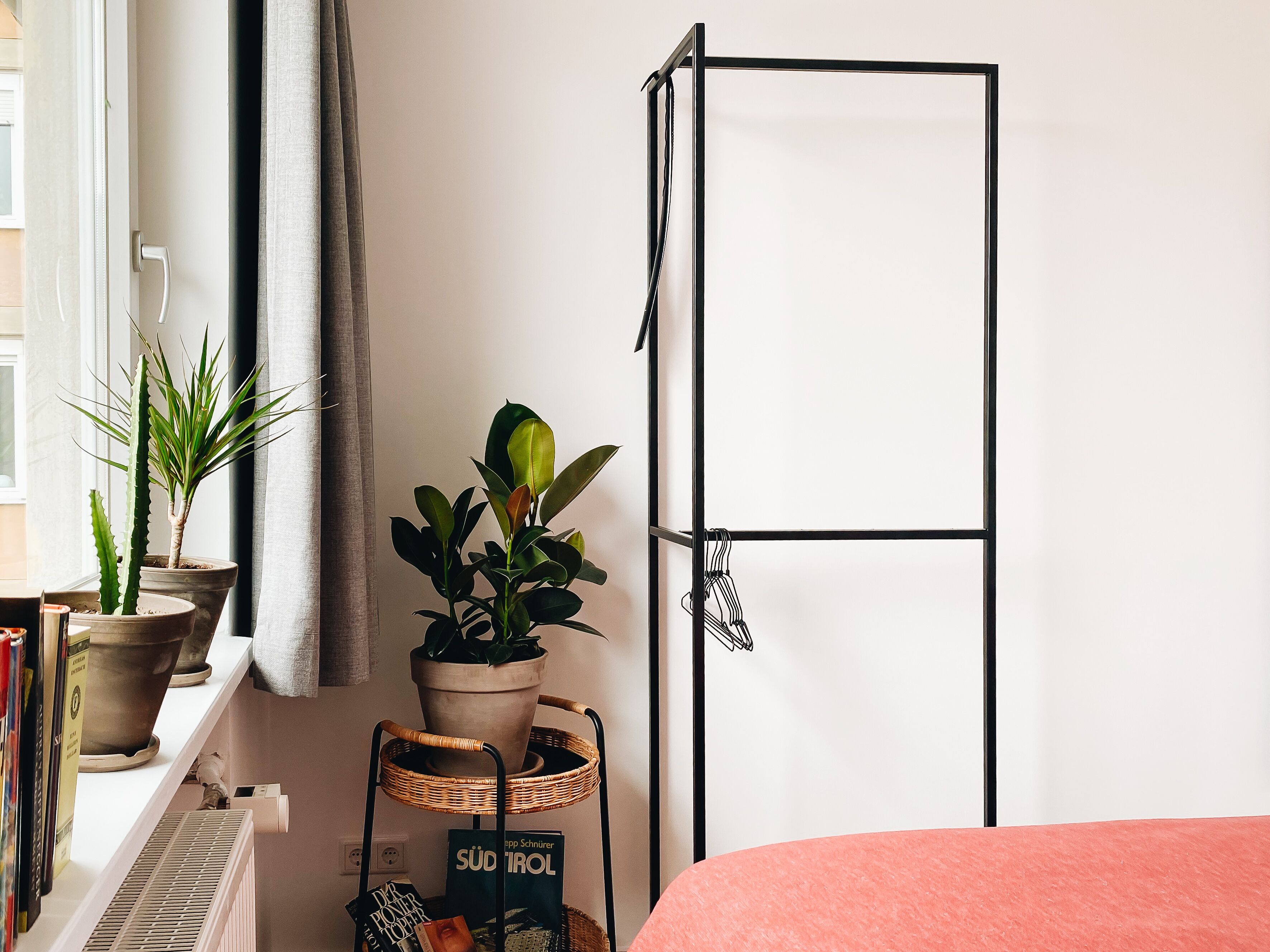 Een kamer met een bed, een radiator, een plank met boeken en een bloemenstandaard met potplanten.