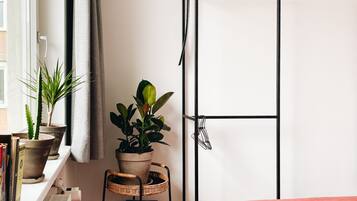 Een kamer met een bed, een radiator, een plank met boeken en een bloemenstandaard met potplanten.