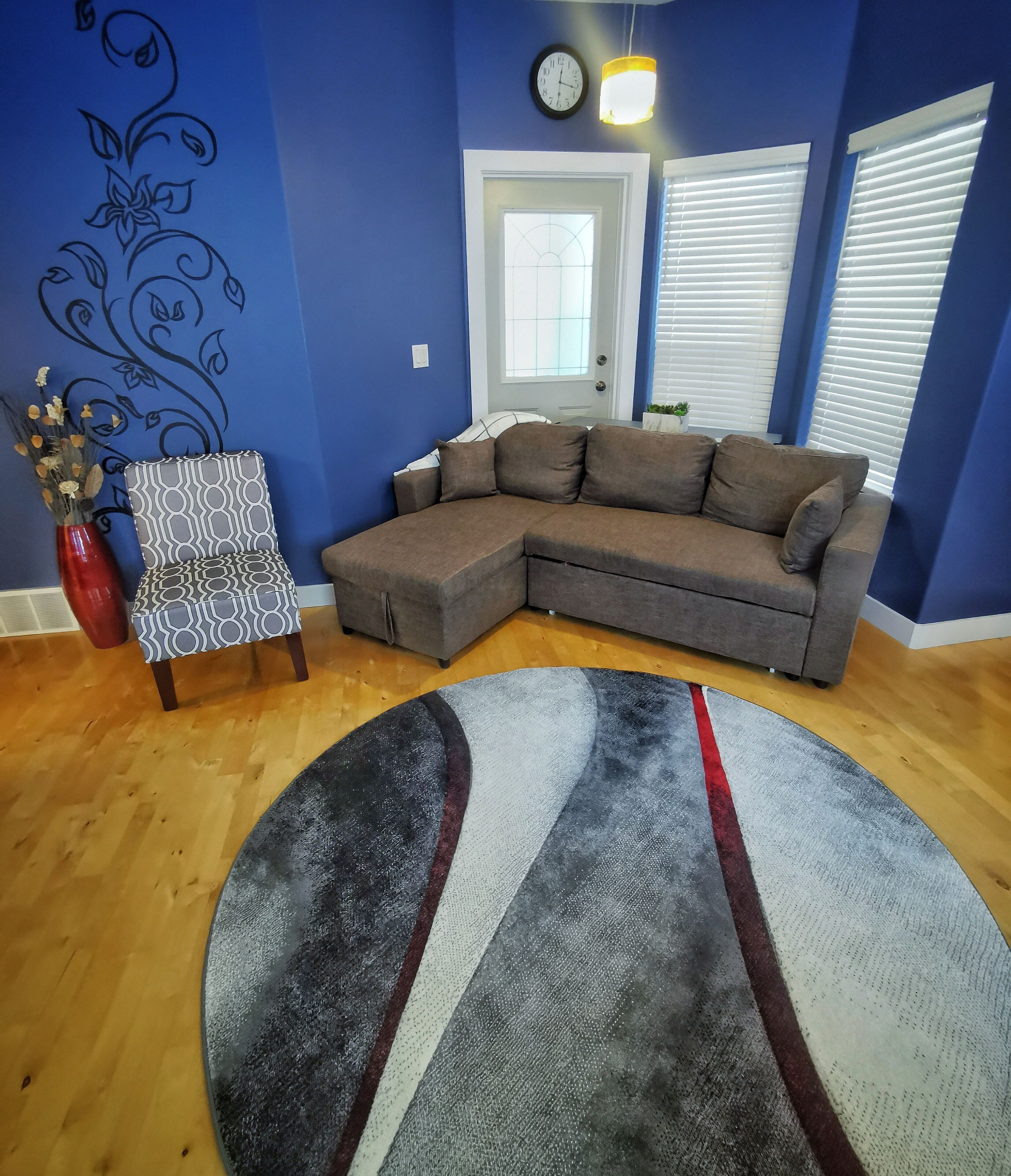 A living room with a sectional sofa, a patterned armchair, a decorative wall, a round rug, and a door with a window.