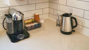 Ett kök med vita kakelväggar, hyllor i trä där vita koppar och glas står, en kaffebryggare, en vattenkokare och en espressomaskin.