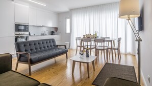 Una sala de estar moderna con sofá, una mesa de centro y un comedor con sillas y una mesa. Hay una zona de cocina con armarios y electrodomésticos blancos.