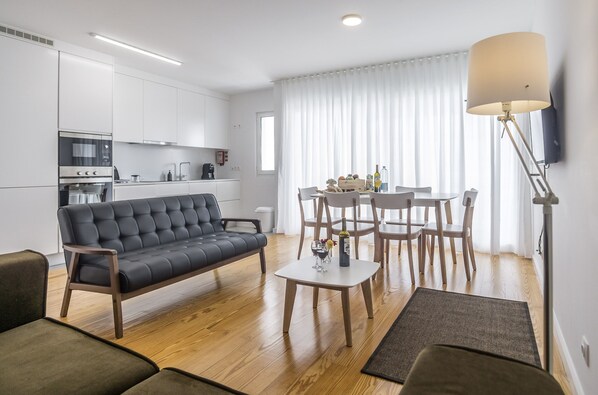Una sala de estar moderna con sofá, una mesa de centro y un comedor con sillas y una mesa. Hay una zona de cocina con armarios y electrodomésticos blancos.