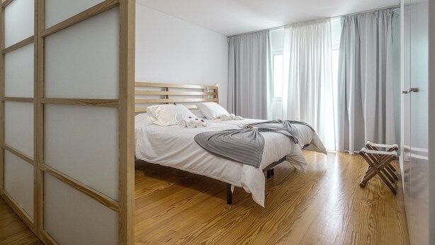 Un dormitorio con cama de madera, ropa de cama blanca y vistas al exterior a través de amplios ventanales.