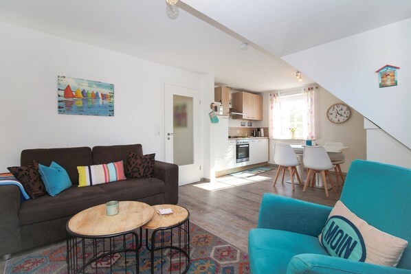Un salon avec un canapé, deux petites tables et un fauteuil turquoise. On aperçoit une cuisine à l’arrière-plan, avec un coin repas.