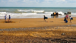 A beach with people walking and carrying surfboards, a boat, and a tractor on the sand.