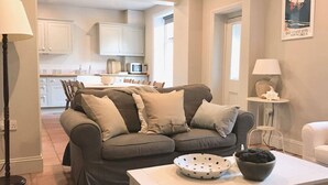 A living room with a grey sofa, a white coffee table, and a lamp. There is a kitchen in the background with white cabinets and a dining area.