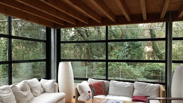 Ein modernes Interieur mit Holzboden, großen Fenstern, einem weißen Sofa, einem Holztisch und Blick auf einen Wald.