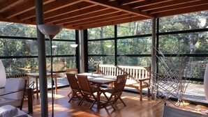 Eine geräumige Terrasse mit Holztisch und -stühlen, große Fenster mit Blick auf die umliegenden Bäume und eine Metallskulptur.