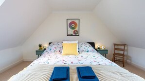 A bedroom with a sloped ceiling, a bed with floral bedding, a yellow pillow, two blue towels, a framed color wheel on the wall, and a small table with a plant and a clock.