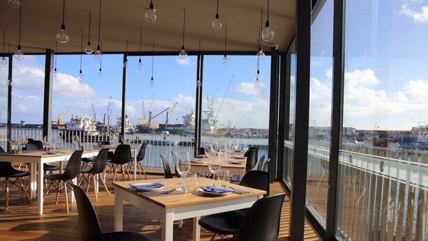 Un espace de restauration en plein air, avec des tables et des chaises, et de grandes fenêtres donnant sur un port de plaisance avec des bateaux et des grues.