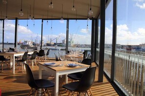 An outdoor dining area with tables and chairs, large windows offering views of a marina with boats and cranes. - Neo City Deluxe Apartment - Ponta Delgada Downtown (Ponta Delgada)