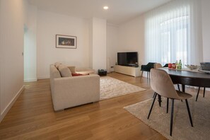 Un salon moderne comprenant un canapé, un téléviseur, une table à manger avec des chaises et une grande fenêtre ornée de rideaux légers.