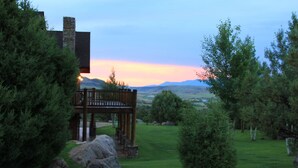 A wooden deck with a stone chimney, surrounded by lush greenery and a view of distant hills. - Elk Run Ranch…Come enjoy the breath taking views of Paradise Valley & YNP (Emigrant)