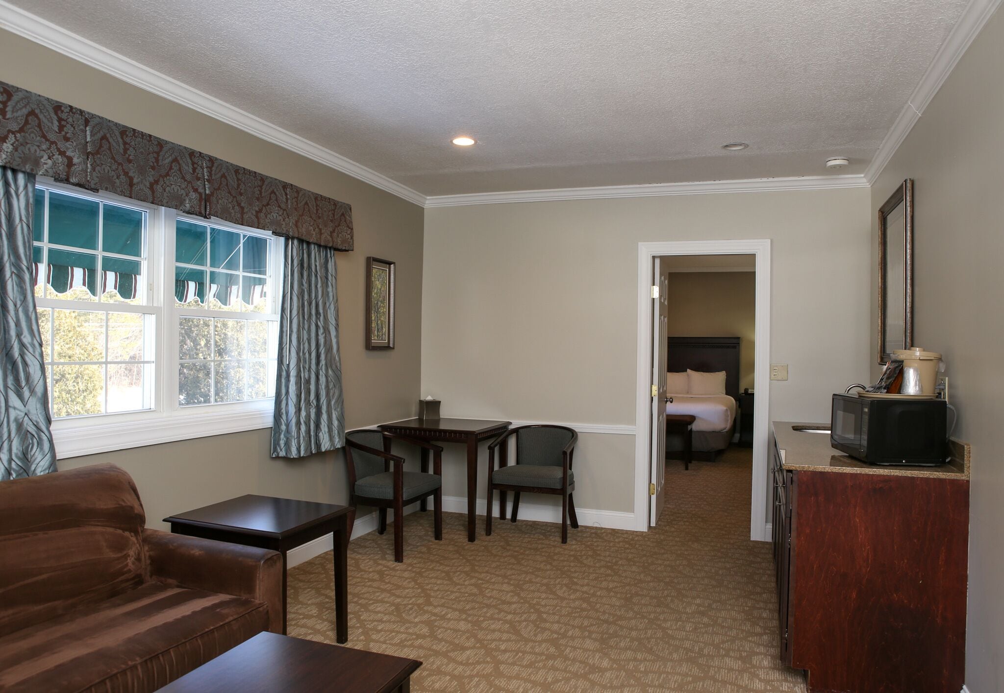 A hotel room with a brown sofa, a small table, two chairs, a kitchenette with a microwave, and a bed in the background.