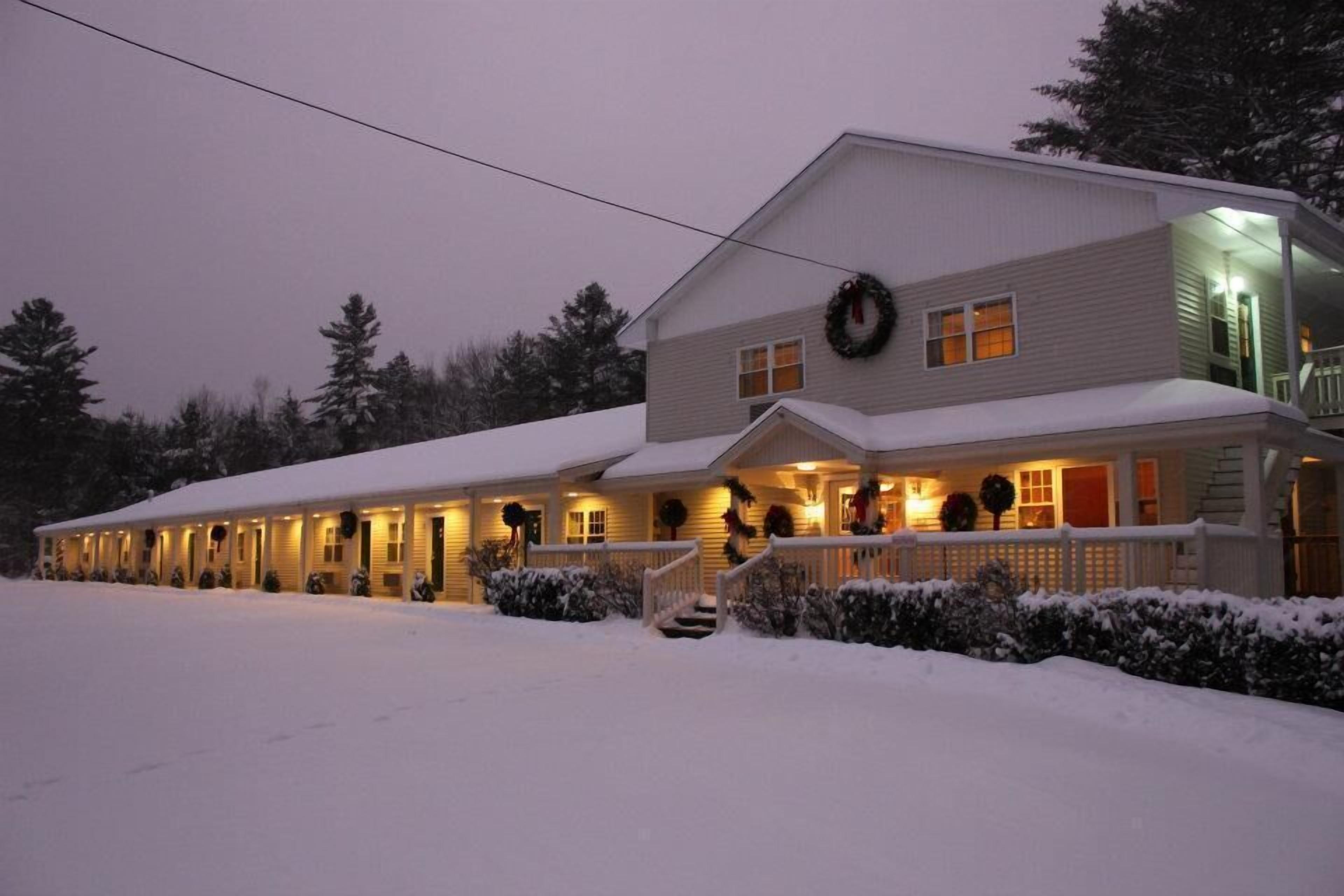 Un chalet à deux étages, dont le toit est recouvert de neige et dont la véranda est illuminée.