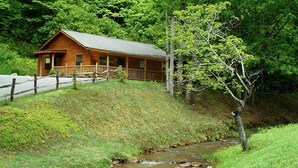 A wooden cabin with a porch, surrounded by lush greenery and a flowing stream. - Log Cabin with Creekside Covered Porch & Outdor Hot Tub! (Bryson City)