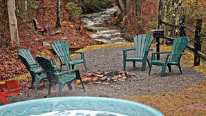 A hot tub surrounded by four green Adirondack chairs, a fire pit, and a wooden fence. - Log Cabin with Creekside Covered Porch & Outdor Hot Tub! (Bryson City)