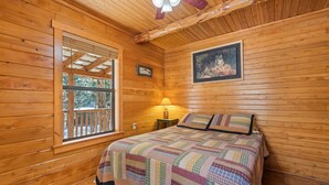 A cozy wooden cabin bedroom with a bed, a window, a lamp, and a painting. - Log Cabin with Creekside Covered Porch & Outdor Hot Tub! (Bryson City)