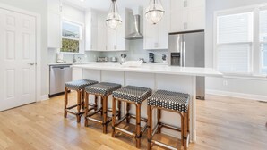 Una cucina moderna con mobili bianchi, un'isola centrale con sgabelli da bar, elettrodomestici in acciaio inossidabile e illuminazione a sospensione.