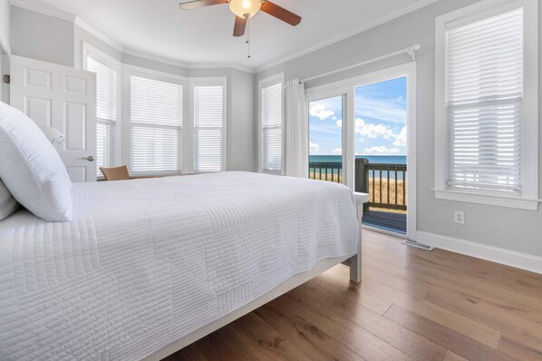 A bedroom with a large bed, a ceiling fan, and a view of the ocean through open windows.