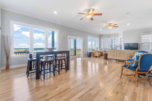 A spacious living area with a wooden floor, a dining table with stools, a sofa, and a TV.