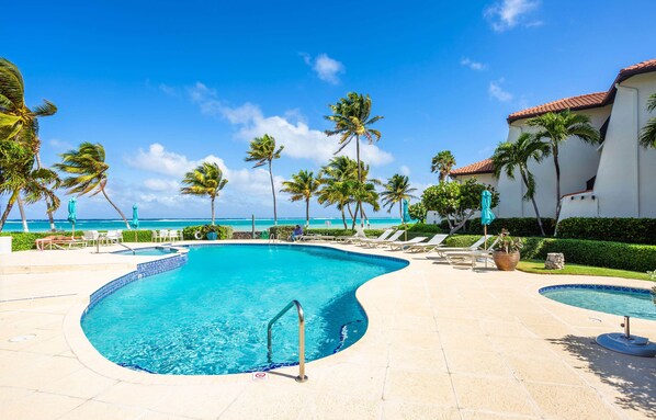 Une piscine à bord incurvé, entourée de chaises longues et de palmiers, avec une vue sur l’océan en arrière-plan.
