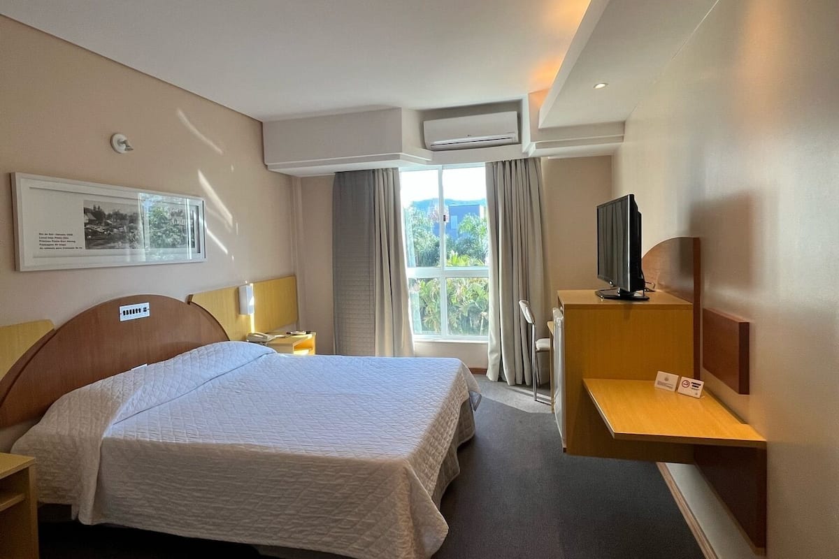 Une chambre d’hôtel comprenant un lit, une table de chevet, une lampe, un appareil de climatisation et une fenêtre avec des rideaux.