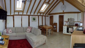 A cozy living room with a sectional sofa, a wooden dining table, and a kitchen area with white cabinets and a red kettle. - Oak Apple - Converted Barn In An Attractive Courtyard On Our Family Stockfarm (Plaitford, Romsey)