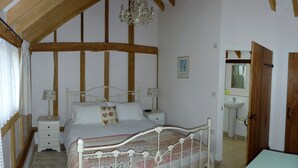 A bedroom with a wooden ceiling, a bed with a floral bedspread, bedside tables with lamps, a hanging chandelier, and a bathroom with a mirror and sink. - Oak Apple - Converted Barn In An Attractive Courtyard On Our Family Stockfarm (Plaitford, Romsey)