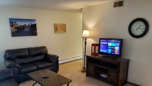 A living room with a black leather sofa, a wooden coffee table, a TV on a wooden stand, a wall clock, and a lamp. - Tomar Park Apt 3 - KP101APT3 (Sioux Falls)